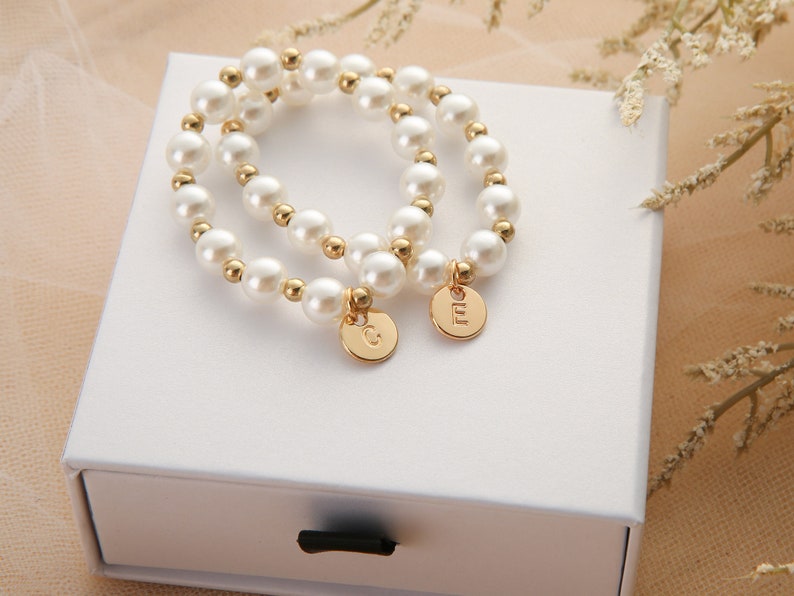 May include: Two pearl bracelets with gold accents and personalized charms. The charms are round and have the letters "G" and "M" engraved on them.