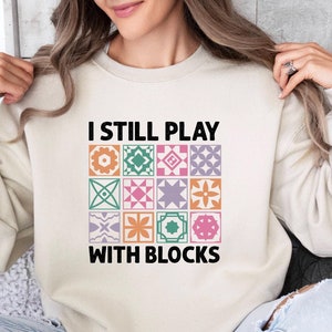May include: A cream-colored sweatshirt with the text "I still play with blocks" in black. The text is surrounded by a colorful pattern of geometric shapes.