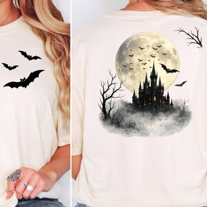May include: A cream-colored t-shirt featuring a small black bat graphic on the front chest area. The back of the shirt displays a large illustration of a dark castle silhouette set against a full moon, surrounded by flying bats and bare trees.