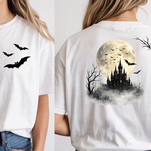 May include: A white short-sleeve t-shirt featuring a small black graphic of three bats on the front chest. The back of the shirt displays a large, detailed illustration of a dark castle silhouette against a full moon with flying bats and bare trees.