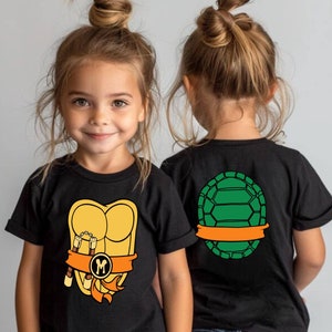 May include: Two children wearing black t-shirts with a Teenage Mutant Ninja Turtles design. One shirt features a green turtle shell on the back, the other shirt features a yellow turtle shell with a brown belt and nunchucks on the front. The shirt also has the letter "M" on the belt.