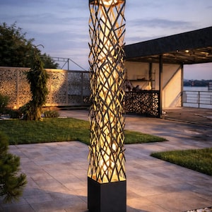 May include: A tall, black outdoor lamp with a geometric, interwoven design. The lamp has a square base and top, with a cylindrical, openwork body that allows light to shine through. The lamp is set on a stone patio with green grass and trees in the background.