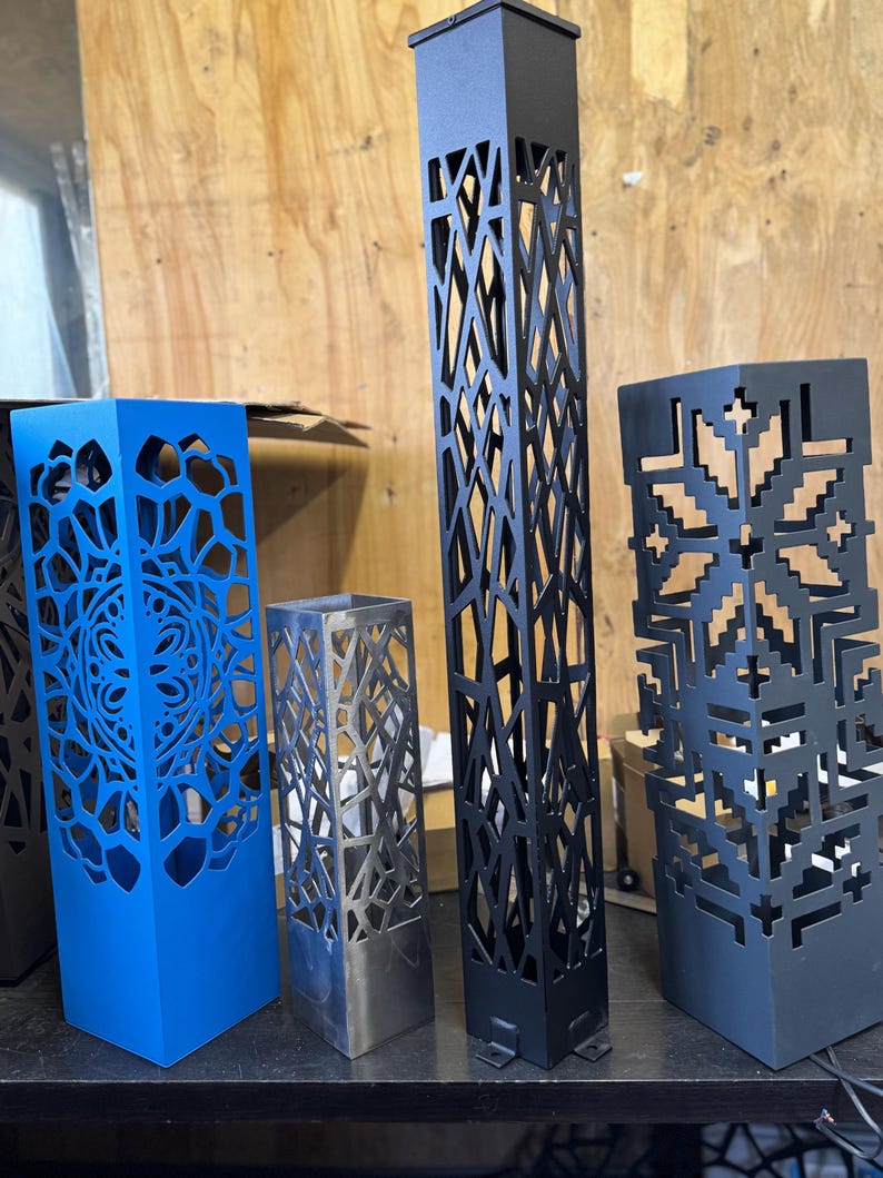 May include: A selection of decorative metal lanterns in various colours and designs. The lanterns are square or rectangular, with intricate cut-out patterns. Colours include blue, black, and silver. The tallest lantern is approximately 91 cm tall.