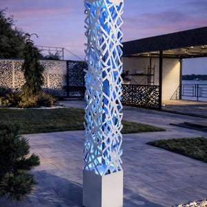 May include: A contemporary outdoor light, featuring a white, geometric design. It has a square base and top, with a latticework pattern in between. The interior emits blue light, creating a striking visual effect. The light is in a garden setting.