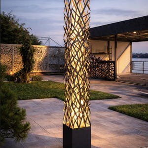 Peut inclure: Lampe d'extérieur haute avec un design géométrique noir. La lampe a une base et un sommet carrés, avec un motif tissé entre les deux. La lampe est éclairée de l'intérieur, projetant une lueur chaleureuse. La lampe est posée sur un patio en pierre. Hauteur environ 180 cm.