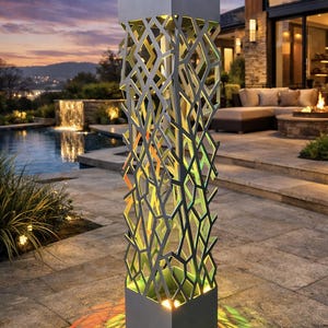 May include: A modern outdoor light fixture with a geometric design. The gray metal structure has a rectangular base and top, with a lattice-like pattern in between. The light projects colorful patterns onto the surrounding patio, with a pool and sunset in the background.
