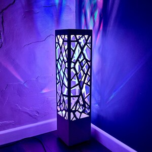 May include: A contemporary floor lamp with a black geometric pattern. The lamp emits a soft, white light from within, contrasting with the dark exterior. The lamp is positioned in a corner, with a black cable running to the floor. The lamp measures approximately 100 cm in height.