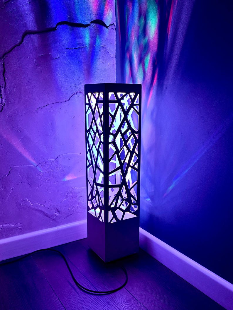 May include: A contemporary floor lamp with a black geometric pattern. The lamp emits a soft, white light from within, contrasting with the dark exterior. The lamp is positioned in a corner, with a black cable running to the floor. The lamp measures approximately 100 cm in height.