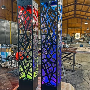 May include: Two black, rectangular, decorative light fixtures with geometric cutouts. One glows red and green, the other blue and purple. The fixtures are on a concrete floor in a workshop.
