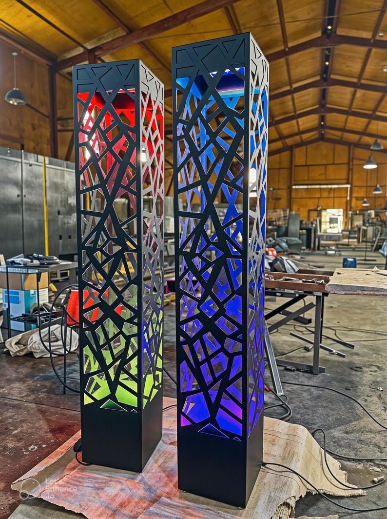 May include: Two black, rectangular, decorative light fixtures with geometric cutouts. One glows red and green, the other blue and purple. The fixtures are on a concrete floor in a workshop.