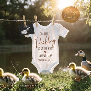 Puede incluir: Un body blanco con el texto "Our little duckling is on the way!" y "Baby Williams September 2025" en texto marrón. El body está colgado de un tendedero con una imagen de ultrasonido borrosa en el fondo. Hay patos en primer plano.