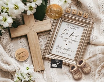 May include: A framed announcement card with a white background and brown text. The text reads "When the time is right, the Lord will make it happen, Isaiah 60:22". Below the text is "Baby Williams, Due August 2025". The card is sitting on a white blanket with a wooden cross and a small wooden circle with the text "Answered Prayer" on it.