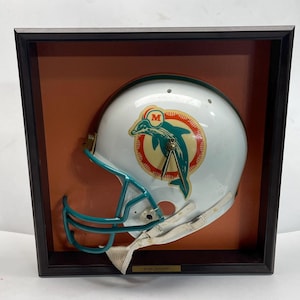 Puede incluir: Un reloj enmarcado con un casco de fútbol americano de los Miami Dolphins. El casco blanco tiene una máscara facial turquesa y un mecanismo de reloj. El marco es negro con un fondo naranja. El texto "MIAMI DOLPHINS" está en una pequeña placa.