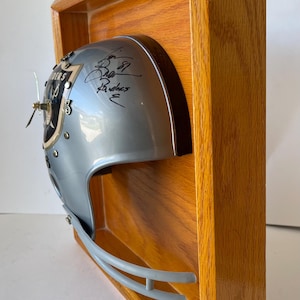 Puede incluir: Un casco de fútbol americano plateado con el logotipo de los Raiders y la firma de un jugador está montado dentro de un marco de madera. El casco tiene una máscara facial gris y está sobre un fondo de madera marrón claro. Un reloj está adjunto.