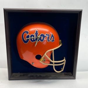 Puede incluir: Un reloj con forma de casco de fútbol naranja con la palabra "Gators" en azul. El reloj está enmarcado en una caja de sombra marrón oscuro con un fondo azul. El casco tiene una máscara facial beige.