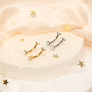 May include: Four curved eyebrow piercing jewelry pieces. Two are gold-toned, and two are silver-toned. Each piece has a small, round, opal-like stone at each end. The jewelry is displayed on a white surface with gold star-shaped confetti.