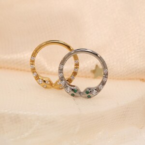 May include: Two hoop earrings, one gold and one silver, each with a snake head design. The gold earring has clear stones, while the silver earring has clear and green stones. The earrings are set against a soft, cream-coloured background.