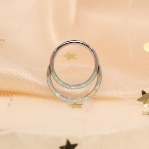 May include: A silver septum ring with a double-band design. The bands feature an iridescent, opalescent material, creating a shimmering effect. The ring is set against a soft, peach-coloured background.