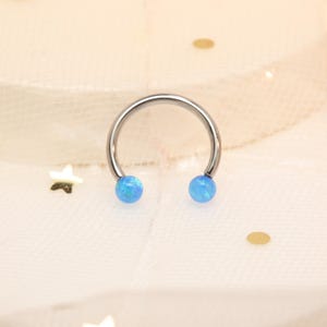 May include: A silver-coloured horseshoe-shaped piercing with two light blue opal-like beads at the ends. The jewellery is set against a soft, off-white background with decorative elements.