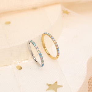 May include: Two delicate rings, one silver and one gold, each adorned with a row of small, iridescent opal-like stones. The rings are set against a soft, neutral background with subtle star-shaped accents, showcasing the jewelry's elegant design.