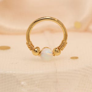 Op de afbeelding: Een goudkleurige septumring met een centrale witte opaalsteen. De ring heeft opgerolde draaddetails aan weerszijden van de opaal en kleine gouden kralen. Ontworpen voor neuspiercings.