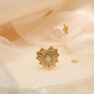 Puede incluir: Un pendiente de oro con un diseño de estrella y una pequeña gema transparente en el centro. El pendiente tiene una superficie texturizada sobre un fondo crema suave.