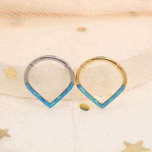 Op de afbeelding: Twee septumringen. Eén is zilverkleurig, de andere goudkleurig. Beide hebben een V-vormige inleg van blauwe opaal. De ringen zijn cirkelvormig met een scharniersluiting. De opaal inleg is een helder, iriserend blauw.