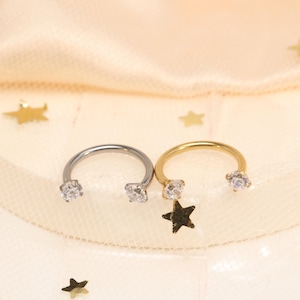 Peut inclure: Deux boucles d'oreilles en forme de fer à cheval. L'une est argentée, l'autre dorée. Chaque boucle d'oreille a une gemme claire et ronde à chaque extrémité. La boucle d'oreille dorée a un petit charme en forme d'étoile sombre. Fond clair et texturé.