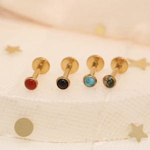 May include: Four gold-colored lip or cartilage studs with different colored gemstones. The gemstones are red, black, turquoise, and green. The studs have a flat back and a small, round gemstone at the front.
