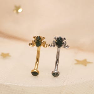 Pode incluir: Dois piercings curvos para sobrancelha, um dourado e outro prateado, cada um com um pingente em forma de sapo com uma pedra verde. Os pingentes de sapo têm pernas minúsculas e uma superfície texturizada. As barras curvas terminam em uma pedra verde combinando.