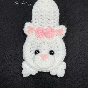 May include: White crocheted cat with a pink bow, black eyes, and a pink nose. The cat is sitting on a black background.
