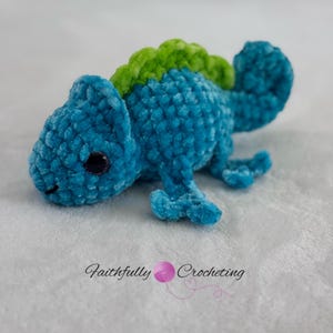 May include: A small, blue, crocheted chameleon plush toy with a bright green dorsal crest and dark, glossy eyes. The textured yarn gives the figure a soft appearance. The text "Faithfully Crocheting" is printed below the plush figure.