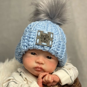 May include: A light blue crocheted baby hat with a gray faux fur pom-pom. The hat has a small tag that says "hello BABY" with an arrow design. The baby is in a woven basket.