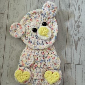 May include: Handmade crochet bear in white with colorful flecks. Features black button eyes, a yellow nose, and yellow heart-shaped feet. The ears and paws are also detailed. The item is placed on a light wood surface. The text "Faithfully Crocheting" is visible.