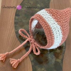 May include: A handmade crochet baby bonnet in a soft peach color with a white stripe. The bonnet has a pointed top and ties with peach-colored yarn. The text "Faithfully Crocheting" is visible in the upper left corner.