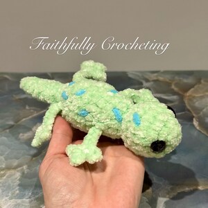May include: A handmade, light green crocheted lizard with blue spots and black button eyes. The plush toy is held in a hand. The text "Faithfully Crocheting" is in the background.