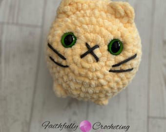 May include: A handmade, crocheted cat toy in a soft, light yellow color. The cat has green eyes, black whiskers, and a stitched 'X' for a mouth. The toy is round and plush, with a small tail. The text "Faithfully Crocheting" is in the background.