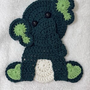 May include: A dark green crocheted elephant applique featuring light green accents on the ears and feet, a white circular belly, and two black plastic eyes. The text Faithfully Crocheting is printed in white script at the bottom of the image.