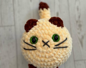 May include: A crocheted cat toy in cream and brown. The cat has green eyes, black whiskers, and a stitched mouth. The text "Faithfully Crocheting" is at the bottom.