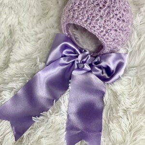 May include: A lavender crocheted baby bonnet with a matching satin ribbon bow. The bonnet is embellished with sequins and has a soft, fluffy interior. The text "Faithfully Crocheting" is in the upper left corner.