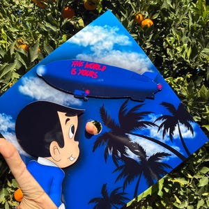 May include: A blue and white artwork featuring a cartoon boy looking at a peach, a blue zeppelin with the text "THE WORLD IS YOURS", and palm trees. The artwork is set against a blue sky background.