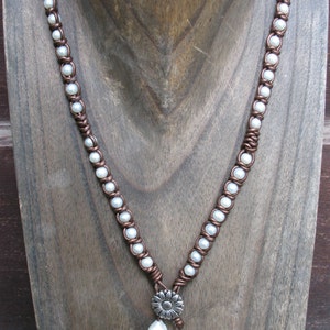 May include: A brown leather necklace with white pearls and a silver flower charm. The necklace is displayed on a wooden mannequin.