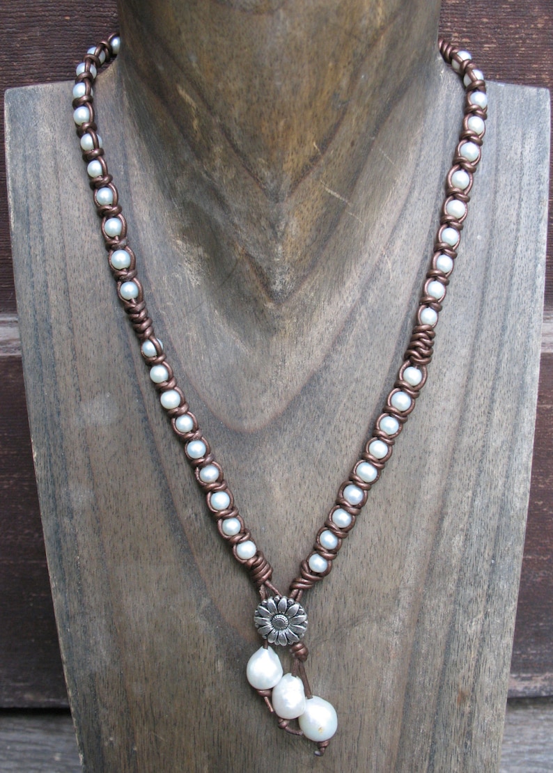 May include: A brown leather necklace with white pearls and a silver flower charm. The necklace is displayed on a wooden mannequin.