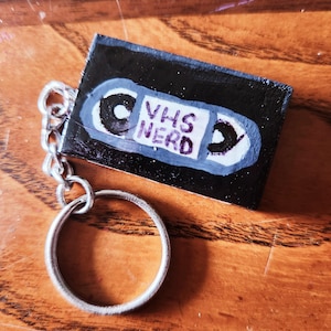May include: A handmade keychain featuring a miniature VHS tape design. The black tape has a white and blue border with the words "VHS NERD" in the center. The keychain is attached to a silver metal chain and ring.