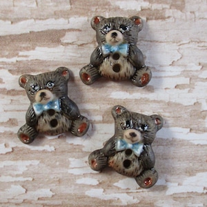 May include: Three ceramic buttons shaped like teddy bears wearing blue bow ties. The bears are brown with white accents and have black eyes.