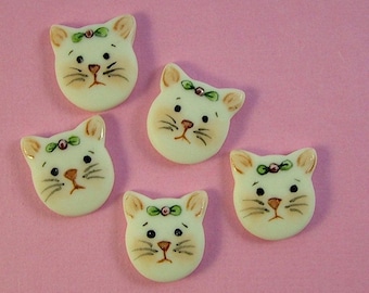 May include: Five white ceramic cat buttons with green and pink painted details. Each button features a cat face with black eyes, a pink nose, and a green bow.