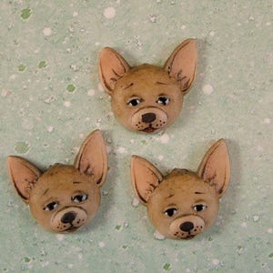 Puede incluir: Tres botones de cerámica con forma de cabeza de perro chihuahua. Los botones son marrones con detalles pintados y tienen un fondo marrón claro.