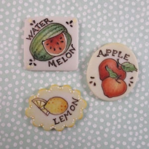 May include: Three decorative ceramic tiles with fruit illustrations. A square tile shows a watermelon with the text "WATER MELON". An oval tile features two apples with the text "APPLE". A scalloped tile displays a lemon with the text "LEMON".