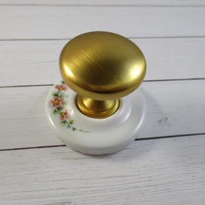 May include: A gold door knob with a white porcelain base featuring a floral design. The base has a pink and green floral pattern.