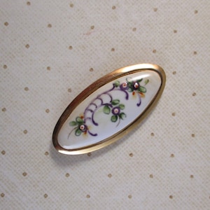May include: An oval-shaped porcelain brooch with a gold-toned metal frame. The porcelain is decorated with a hand-painted floral design in shades of purple, green, and white.